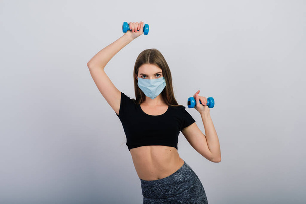 Young woman in a protective mask with dumbbells on a white background. Quarantined sports - Photo, Image