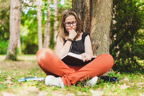 Student czyta książkę w parku. Nastolatek w czerwonych okularach studiuje na zewnątrz, opierając się o siedzenie na drzewie - Zdjęcie, obraz