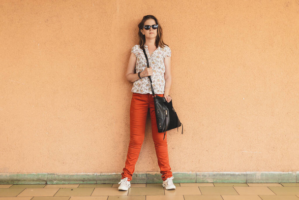 Beautiful woman model leaning against an orange wall outside. Pretty girl with brown hair wearing sunglasses and a casual outfit daydreaming while looking away in the distance - Photo, Image