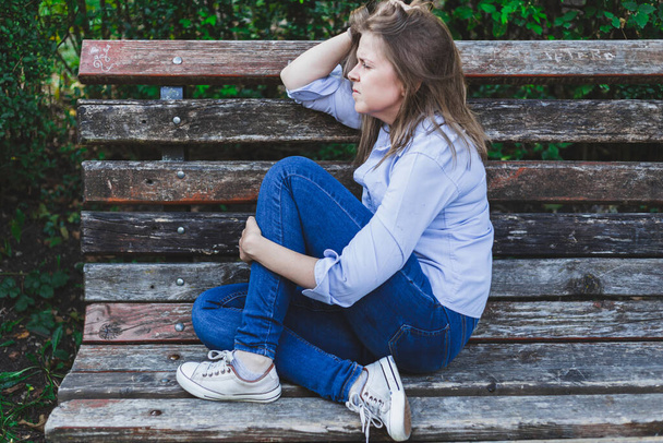 Sad and pensive woman sitting on a bench in the park. Casually dressed woman wearing jeans and shirt feeling depressed while being alone outdoors. Cute female feeling bad and lonely - Foto, Bild