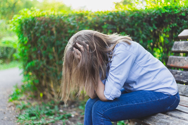 Sad and depressed young woman holding her head with both hands while looking down outdoors. Anxious female worker wearing jeans and blue shirt having panic attack in the park while sitting on a bench - Photo, Image