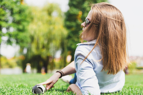 Jonge gelukkige vrouw die naar muziek luistert op een kleine draagbare luidspreker terwijl ze buiten in het gras ligt. Vrijetijds gekleed meisje met bruin haar in een zonnebril ontspannen en genieten van vrije tijd in het park - Foto, afbeelding