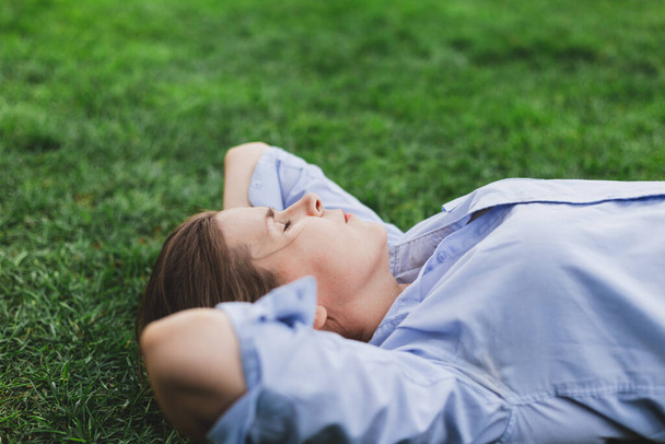 Çimlerin üzerinde yatarken dışarıda uyuyan yorgun genç bir kadın. Doğada kestirirken enerjiden boşalan sevimli kadın işçi. Kahverengi saçlı kız iki elini başının altında tutuyor. Parkta gözleri kapalı rahatlıyor. - Fotoğraf, Görsel