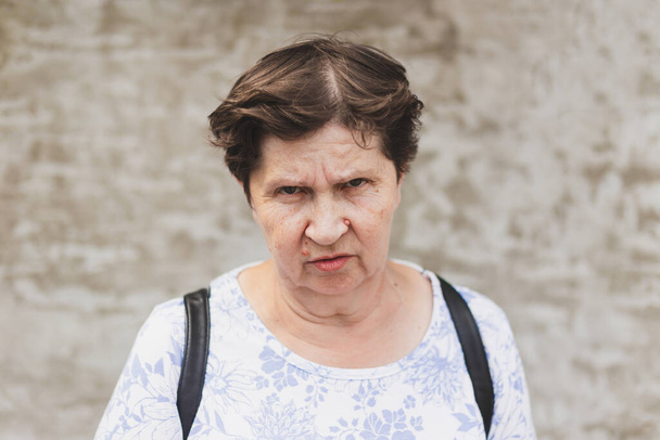 怒りの表情をした先輩女性の肖像画。通りに一人で立って老婦人を動揺させます。不幸なでこぼこの高齢者の距離でクレイジーに見える - 写真・画像