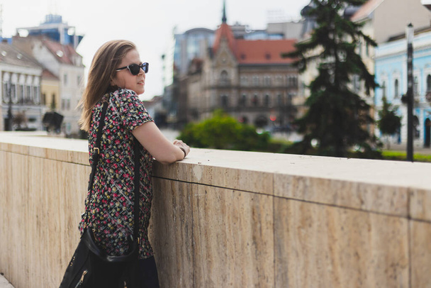 Woman admiring cityscape while standing on a stone riling. Beautiful young female wearing sunglasses relaxing while looking in the distance outdoors - Фото, зображення