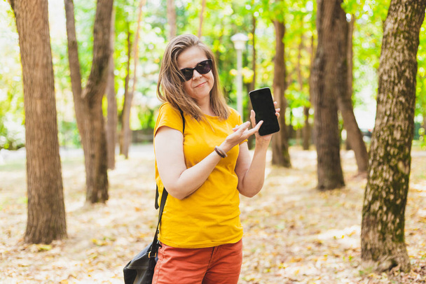 Θλιβερή νεαρή γυναίκα που δείχνει σε ένα μαύρο κινητό τηλέφωνο να στέκεται έξω μια καλοκαιρινή μέρα. Δυστυχισμένη έφηβη που φορούσε γυαλιά ηλίου κρατώντας ένα μικρό μαραφέτι στο πάρκο. Έκπληκτη γυναίκα με σύγχρονη τεχνολογία - Φωτογραφία, εικόνα