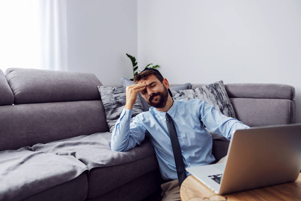 Handsome bearded businessman dressed elegant sitting on the floor at home and holding hand on head. There is a laptop on the table. - Фото, изображение