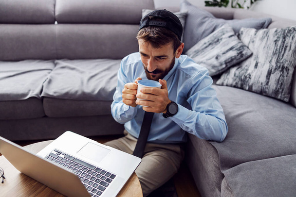 Молодой бизнесмен, одетый деловым случайно сидеть дома на полу, пить кофе и смотреть на ноутбук
. - Фото, изображение