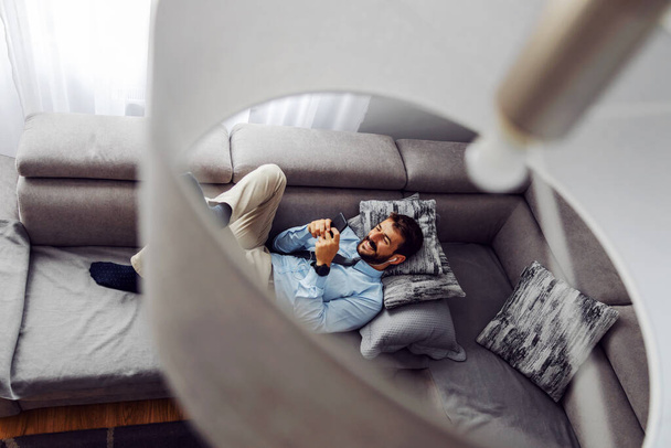 Giovane uomo d'affari attraente sdraiato sul divano a casa, rilassante e utilizzando lo smartphone. - Foto, immagini