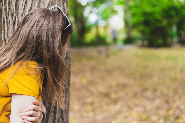 Bang en paranoïde jonge vrouw verstopt zich achter een boom in het park. Zenuwachtig en achterdochtig meisje dat de omgeving vanuit een veilige plek observeert - Foto, afbeelding
