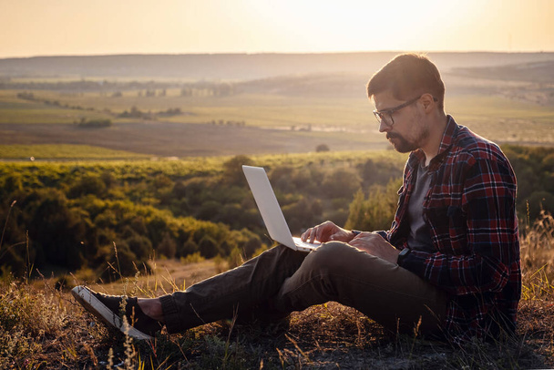 man met laptop zittend aan de rand van een berg met een prachtig uitzicht op de vallei. Mobiel netwerk broaband dekking concept. 5G. - Foto, afbeelding