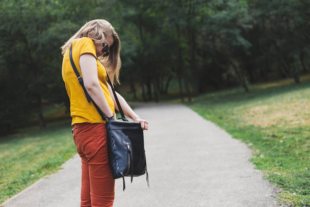 Young woman searching for her lost keys into her purse outdoors. Casually dressed girl with brown hair looking nervously in her black leather bag while walking in the park - Foto, afbeelding