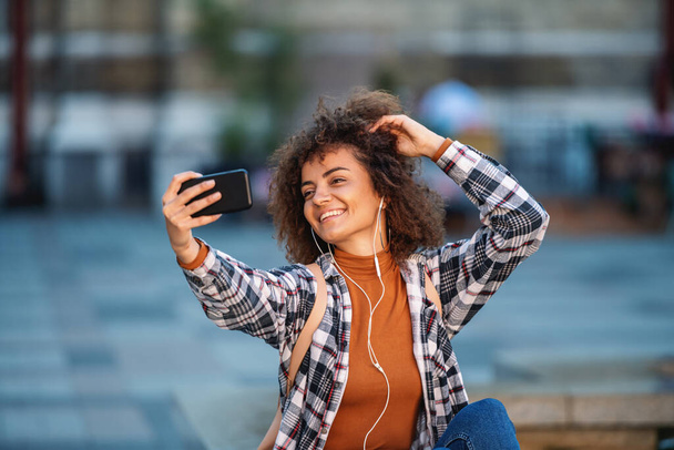 Молода приваблива змішана дівчина, що сидить на фонтані, слухає музику і приймає селфі для соціальних медіа
. - Фото, зображення