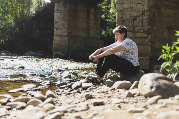 Sad senior woman sitting alone near a river on a summer day. Depressed and lonely old lady looking in the distance in nature. Concept image for abandon and loneliness - Photo, Image