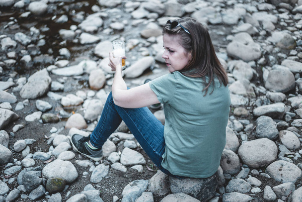 Nainen, jolla on olutta ja joka istuu kivillä joen reunalla. Yksinäinen matkustaja nauttimassa hetkestä pitäen lasillisen alkoholia - Valokuva, kuva