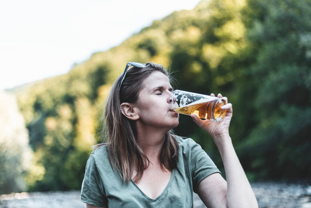 Beautiful young woman drinking beer while relaxing in nature on a summer day. Happy casually dressed girl enjoying cold drink in nature on a hot day - Zdjęcie, obraz