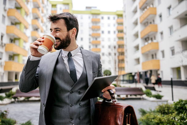Junger lächelnder Geschäftsmann, der Kaffee trinkt und zur Arbeit geht. Im Hintergrund sind Gebäude. - Foto, Bild