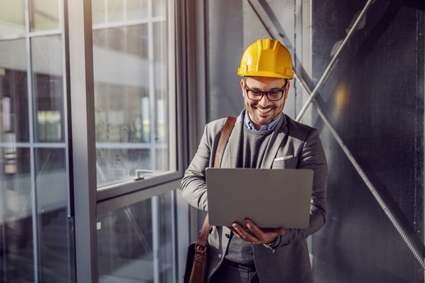 Młody uśmiechnięty architekt stojący w pobliżu okna w budynku w procesie budowy i przeglądający plany na laptopie. - Zdjęcie, obraz
