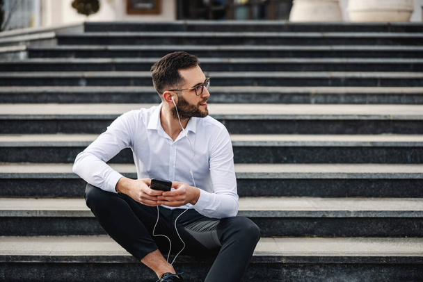 Νεαρός ελκυστικός γενειοφόρος επιχειρηματίας κάθεται στις σκάλες μπροστά από την εταιρεία του, κρατώντας έξυπνο τηλέφωνο και ακούγοντας μουσική. - Φωτογραφία, εικόνα