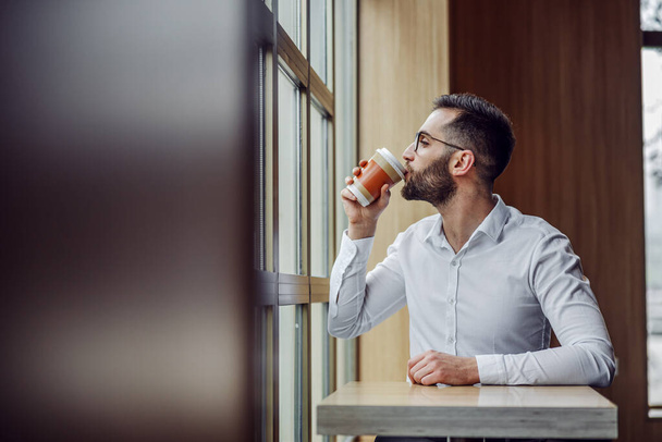 Молодий красивий бородатий елегантний чоловік сидить у кафетерії і п'є свіжу ранкову каву з одноразової чашки
. - Фото, зображення