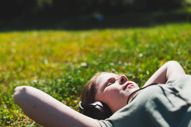Beautiful woman relaxing in nature while listening to music. Cute girl with brown hair wearing big headphones enjoying the sun while lying in the grass on a bright summer day - Fotografie, Obrázek