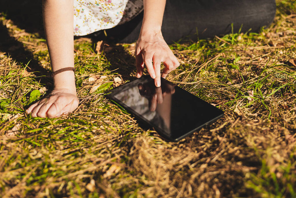 Young woman using a tablet while sitting in the grass outdoors. Female writing on a touchscreen gadget in the park. Person with digital technology - Photo, image