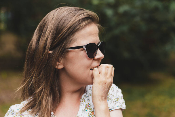 Zenuwachtige jonge vrouw die op haar nagels bijt. Gestresst persoon met een zonnebril met een onhygiënische gewoonte. Meisje houdt vingers in haar mond - Foto, afbeelding
