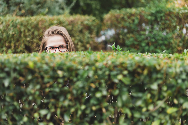 Femme effrayée se cachant derrière un buisson pendant la journée time.Cute fille aux cheveux bruns portant des lunettes de vue ont peur debout seul dans le parc - Photo, image