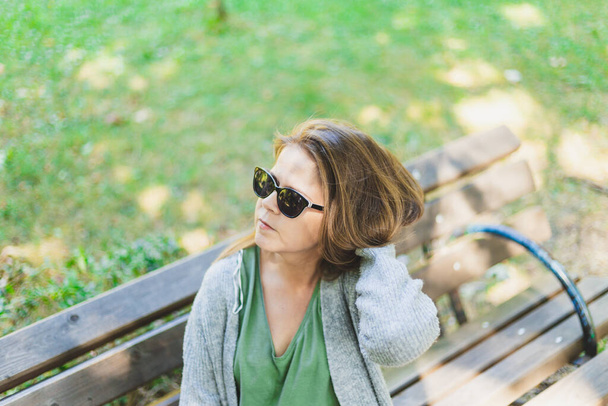 Mulher a arranjar cabelo lá fora. Menina vestindo óculos de sol e sentado em um banco fora ajustando seu estilo de cabelo
 - Foto, Imagem
