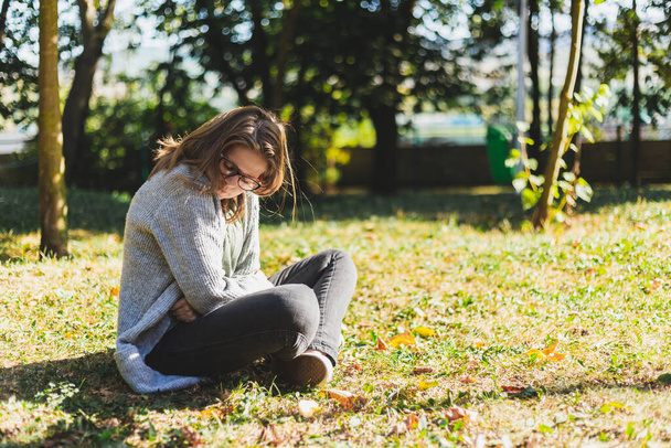 Sad young woman suffering from period pain sitting down outdoors. Beautiful girl with brown hair wearing jeans having menstruation cramps while holding her belly in the park - Foto, Imagen