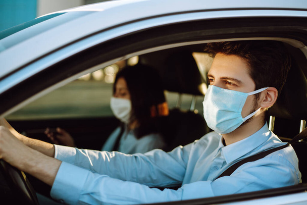 Young man and woman  driving in a car during coronavirus quarantine in medical masks. Safe taxi, protection from the virus. Covid-2019. - Valokuva, kuva