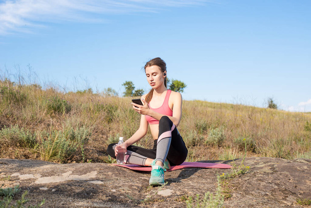Mujer caucásica joven sentada en una esterilla de yoga con botella de agua, mirando el teléfono celular con auriculares, relajándose con la música después de ejercicios deportivos
 - Foto, Imagen