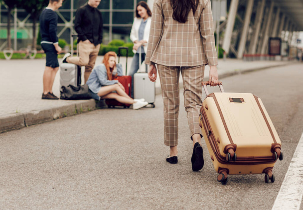友人のグループに近づいて荷物を持つ女性の背面図。人々はアスファルトの飛行を待って座っている。友人との旅行、休暇 - 写真・画像