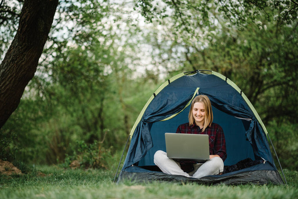 Woman working on laptop in tent in nature. Young freelancer sitting in camp. Relaxing in camping site in forest, meadow. Remote work, outdoor activity in summer. Happy girl relaxing, work on vacation. - Foto, Imagen