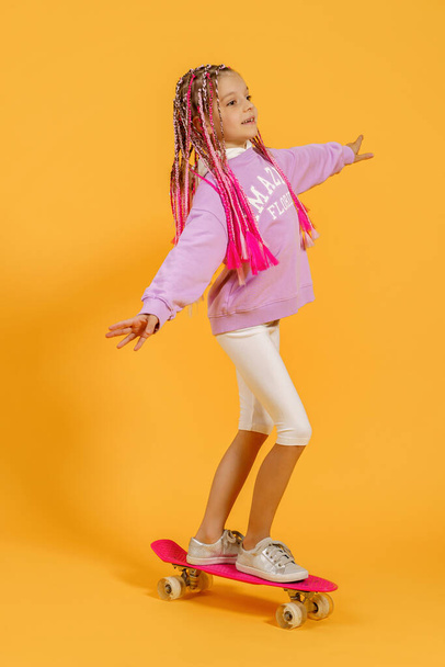 Active young girl in pink shirt and white shorts holding skateboard over yellow background - Fotoğraf, Görsel
