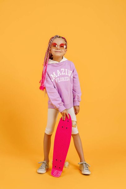 Active young girl in pink shirt and white shorts holding skateboard over yellow background - Φωτογραφία, εικόνα