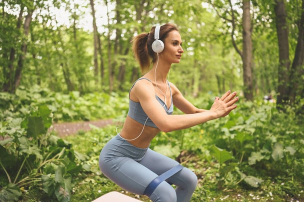 Junge glückliche schöne schlanke Frau in blauer Sportbekleidung mit großen weißen Kopfhörern beim Kniebeugen mit Fitnessgummi im sommergrünen Park im Freien. Mädchen mit perfektem Körper, Sport Yoga - Foto, Bild