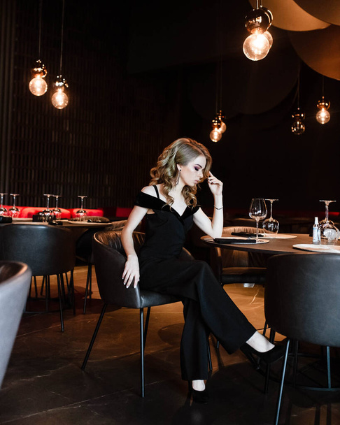 Beautiful blonde, young girl. Portrait of a young girl in a restaurant. Young girl in a restaurant on a black background. Portrait of a girl in a black dress. Young girl. - Photo, Image