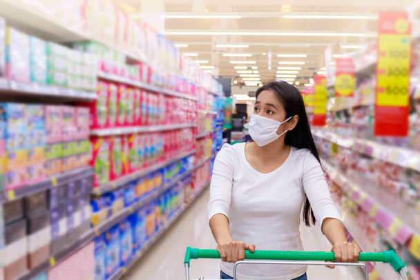 Азиатка носит маску для лица толкать корзину в супермаркете. Девушка ищет продукты, чтобы купить что-нибудь. Во время коронавирусного кризиса или вспышки ковида19. Женщины в защитной маске для лица новая нормальная концепция
. - Фото, изображение
