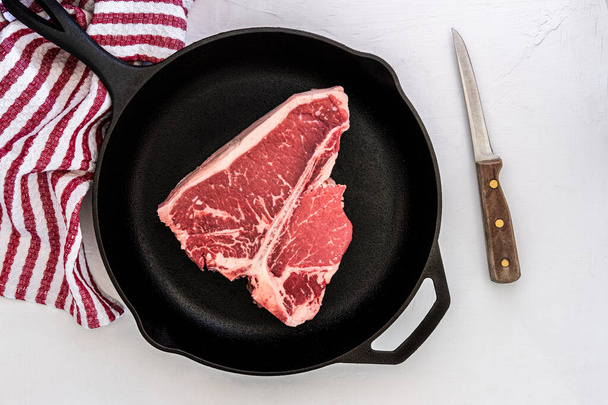 Foto eines T-Bone Choice Steaks in einer gusseisernen Pfanne mit einem Knochenmesser und Handtuch im Hintergrund - Foto, Bild