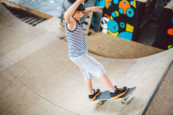 Детский скейтбордист во время обучения трюкам на рампе в городском скейт-парке. Мальчик в спортивном шлеме едет на конькобежной доске в спортивном зале. Активные экстремальные дети проводят время. парень катается на скейтборде
 - Фото, изображение