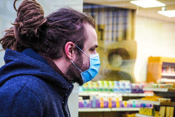 Handsome young European man with a medical face mask on looking inside the pharmacy - Photo, Image