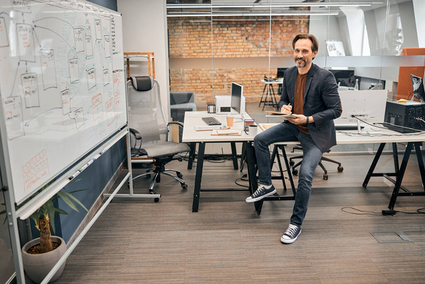 Smiling leader at company wearing jeans and jacket sitting on the table - Photo, image