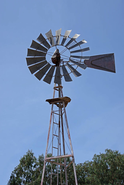 Vista de ángulo bajo de un viejo molino de viento de equipo de granja bajo un cielo azul brillante
 - Foto, Imagen