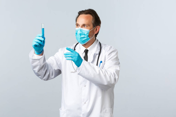 Covid-19, preventing virus, healthcare workers and vaccination concept. Excited doctor in clinic lab developed coronavirus vaccine, wear medical mask and gloves, looking thrilled at test-tube - Foto, Imagen