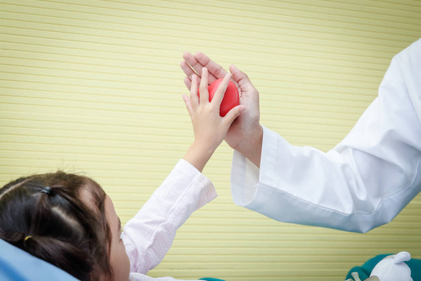 Μια γυναίκα γιατρός με μια κόκκινη μπάλα στην καρδιά Αγγίζοντας τα χέρια με ένα μικρό κορίτσι που είναι άρρωστο. Η έννοια της θεραπείας των παιδικών καρδιακών παθήσεων. - Φωτογραφία, εικόνα