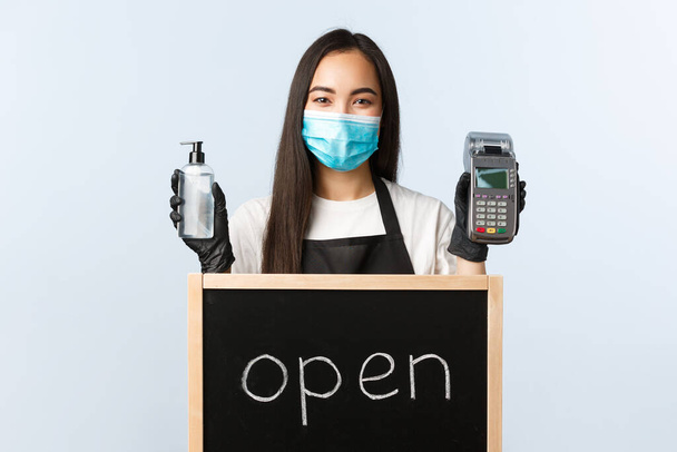 Small business, covid-19 pandemic, preventing virus and contactless orders concept. Cute asian female barista or waitress with we are open sign, showing hand sanitizer and paying terminal - Photo, Image
