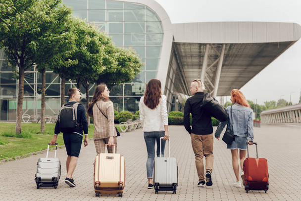 Vista trasera de los jóvenes positivos con equipaje al aire libre. Amigos van al aeropuerto para volar de vacaciones
 - Foto, Imagen