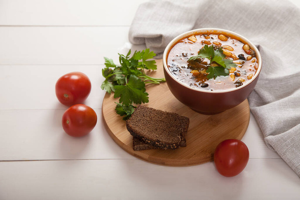 Bean soup with vegetables, olives, cheese and fresh herbs in a ceramic bowl, next to rye bread, tomatoes on a wooden stand. White table, linen napkin - Фото, зображення