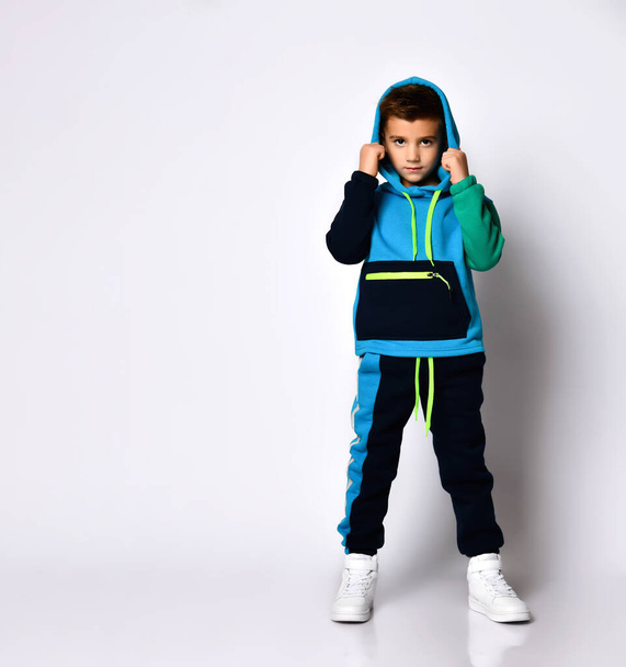 Little child in colorful sport suit and sneakers. He has put his hood on while posing isolated on white studio background - Foto, Imagem